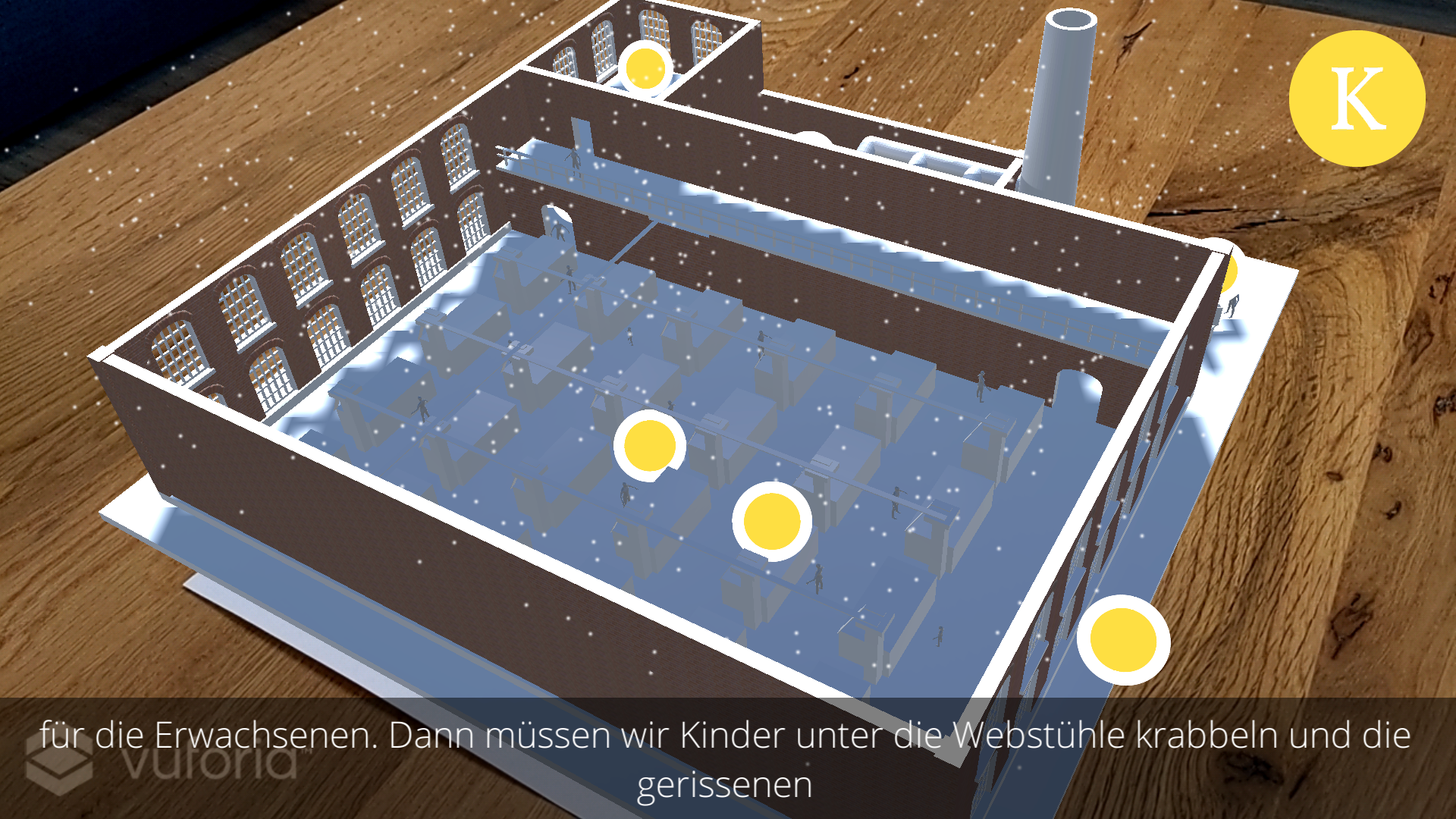 Screenshot of yARn that shows a 3D rendered factory projected on a wooden table from the perspective of the child. There are several yellow colored points of interest inside the factory. At the bottom of the screen subtitles are displayed.